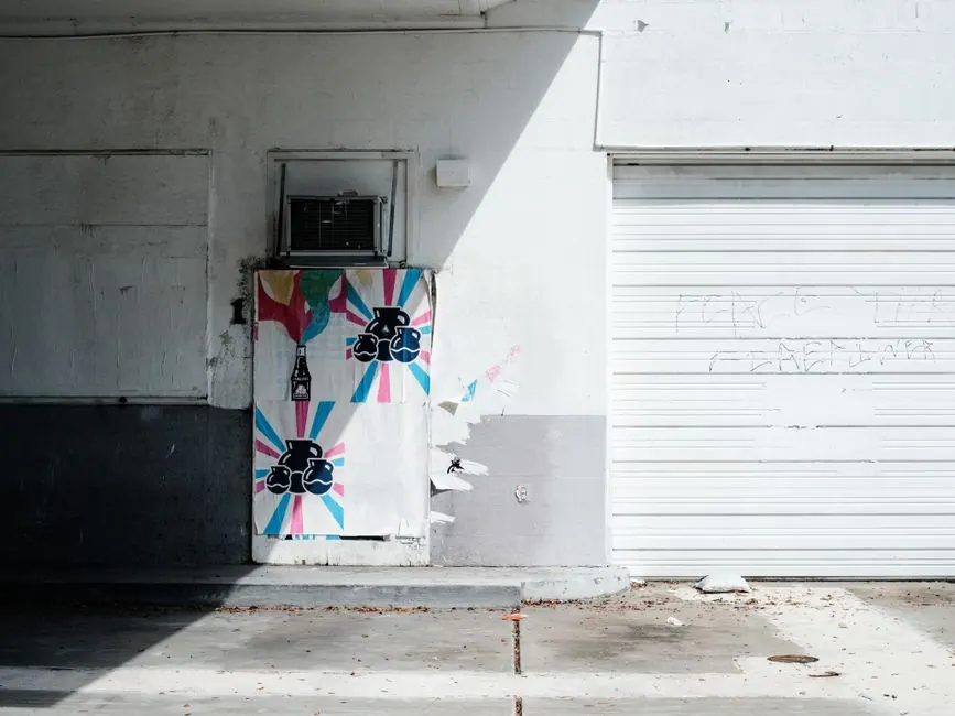 Street art on a sunlit wall with a white garage door and colorful posters.